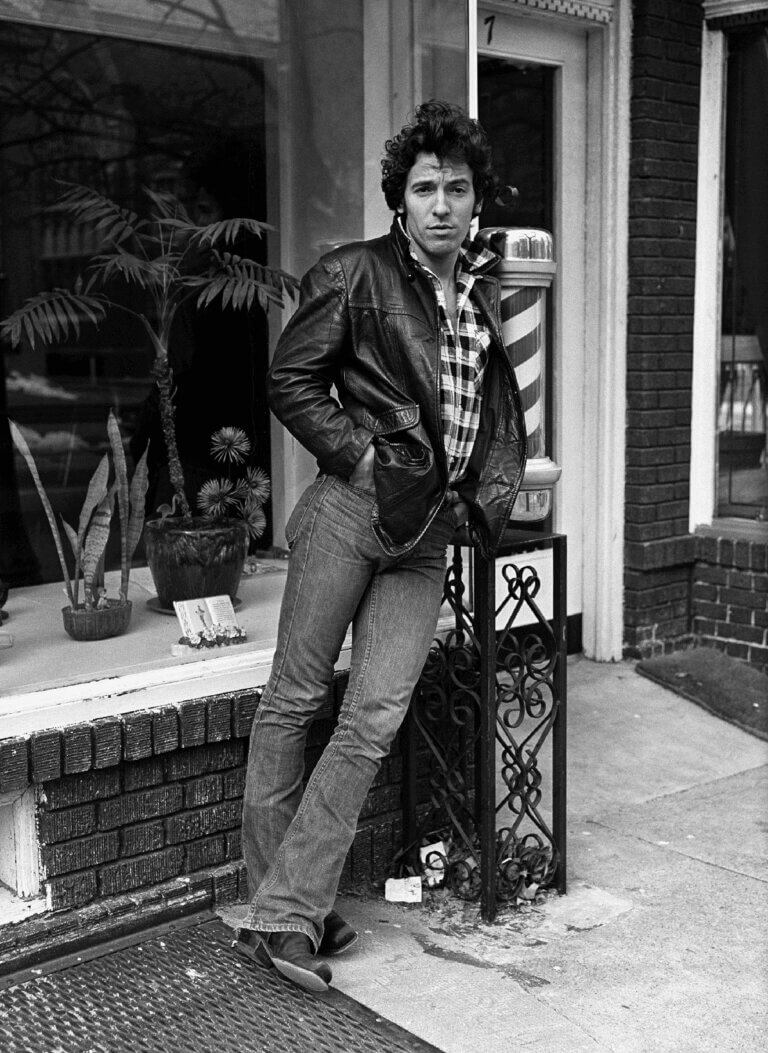 Bruce Springsteen in a leather jacket leaning on a railing outside a storefront, black-and-white photo
