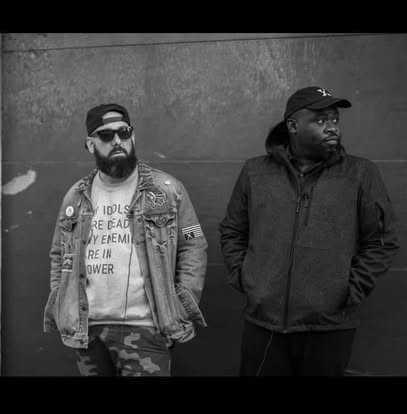 2 men leaning against a wall with hands in pockets wearing caps on heads. one is wearing dark glasses. both are wearing jackets. one man has a beard.
