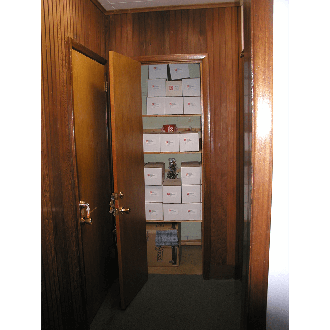 A small storage room inside the old Bruce Springsteen Archives, viewed through a partially open wooden door. The shelves are lined with white archival boxes labeled with red logos, and the walls and door frames are covered in dark wood paneling.
