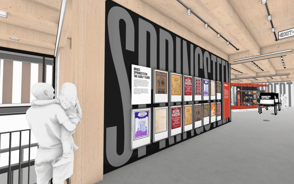 A modern museum exhibit featuring large "SPRINGSTEEN" text on walls, with framed posters displayed. A person holds a child, observing the display.