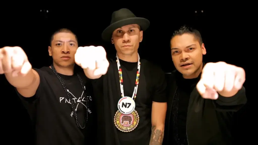 Three individuals facing forward, fist-bumping the camera. The central figure wears a fedora and beaded necklace. The image exudes confidence and unity.