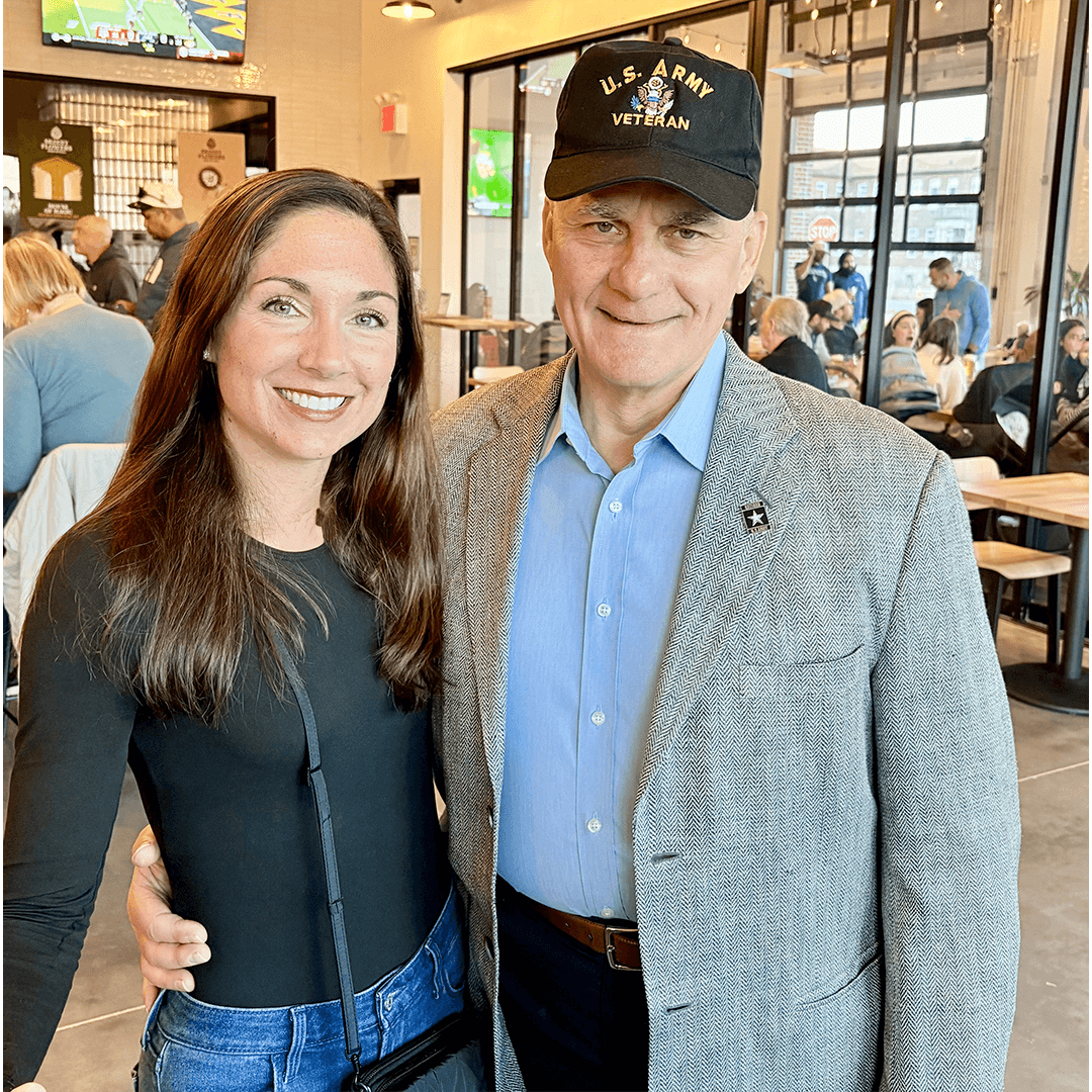 A woman and a man stand smiling in a busy cafe. The man wears a "U.S. Army Veteran" cap and a gray jacket. The scene is warm and social.