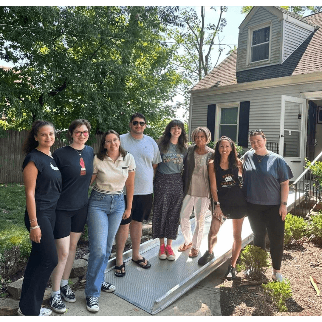 A group of eight people stands together outside a house on a sunny day, smiling warmly. Trees and the house create a welcoming, friendly atmosphere.