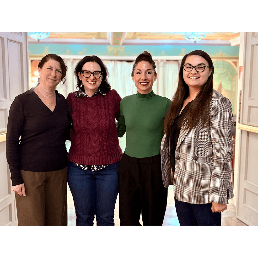 Four women stand together, smiling warmly. They are in a room with elegant decor, featuring soft colors and warm lighting, exuding a friendly atmosphere.