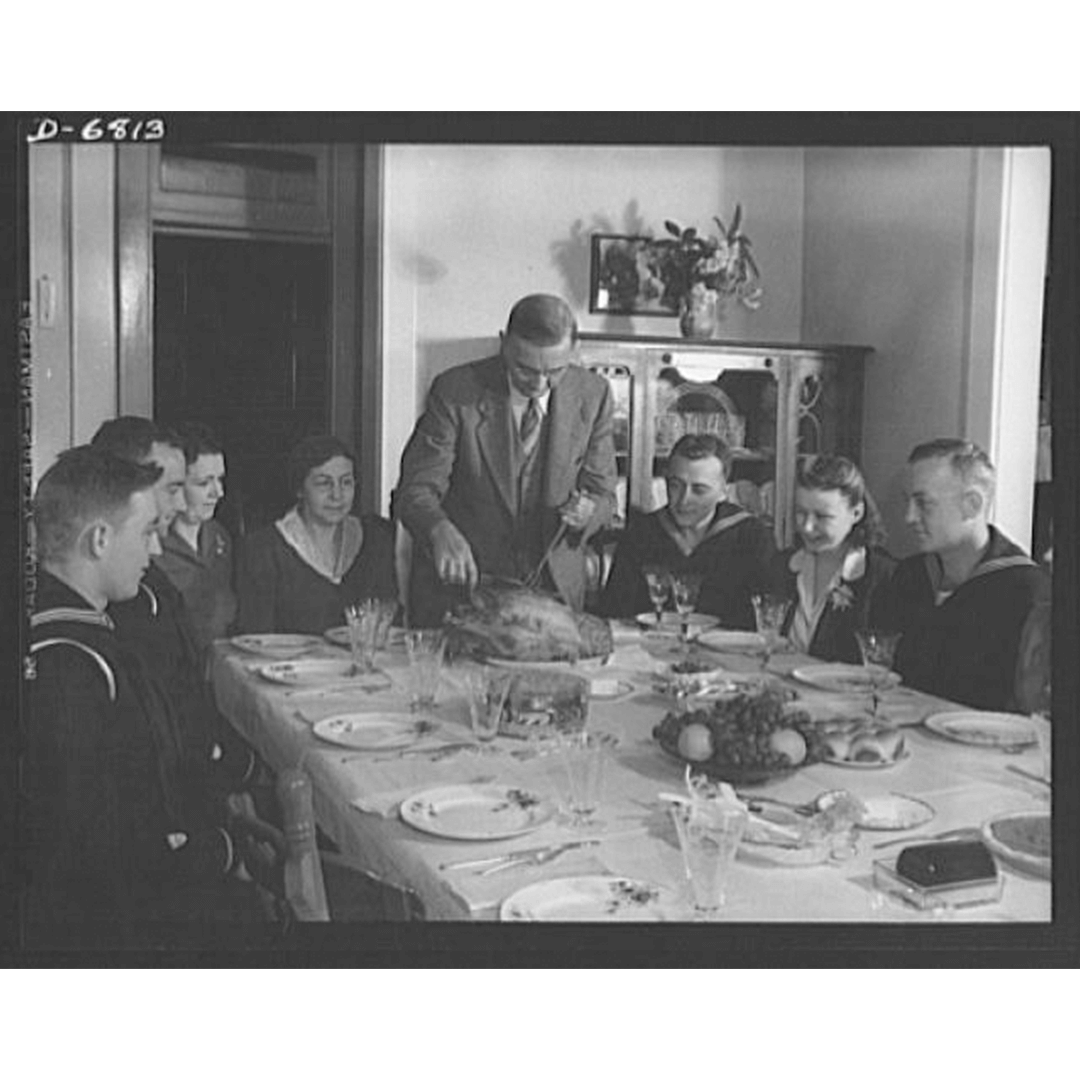 A group of people gather around a dining table for a festive meal. A man carves a turkey while others, including uniformed individuals, watch. The room has warm lighting and a cozy atmosphere.