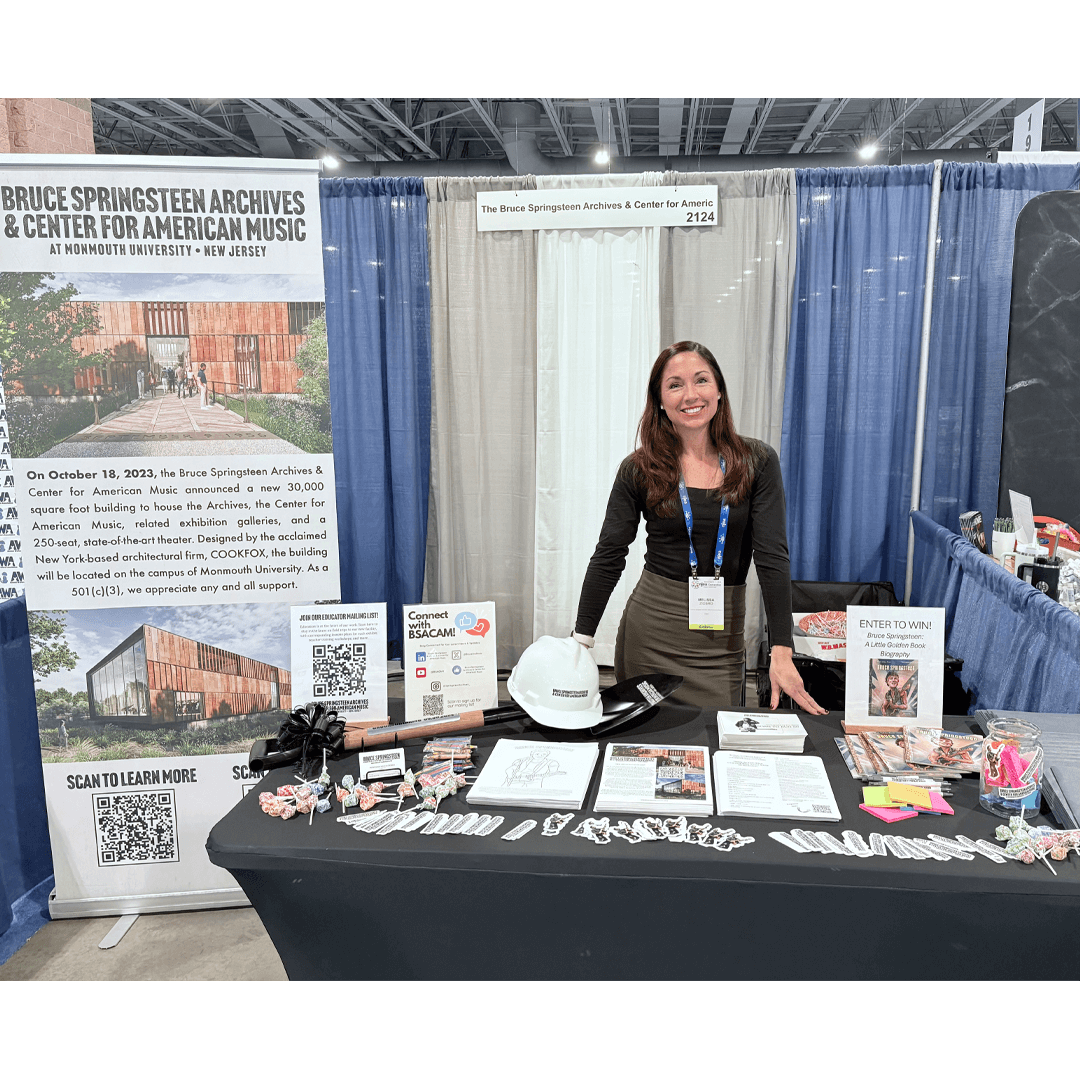A woman stands smiling behind a booth for the Bruce Springsteen Archives & Center for American Music. The table displays brochures, QR codes, and promotional items, including a branded hard hat and small toys. The backdrop features informational posters about the center. The setting conveys an inviting and informative atmosphere at a convention.