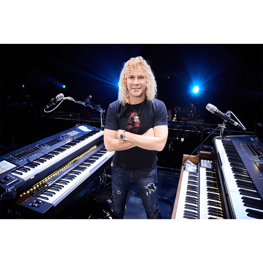 A person with curly blond hair smiles while standing between two keyboards on a dark stage. Bright lights shine in the background, creating a vibrant atmosphere.
