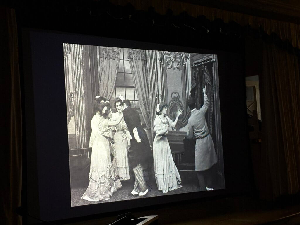 Black and white photo of a staged theatrical scene. Five actors in period costumes; women in elegant dresses and a man pulling back curtains. Victorian setting.