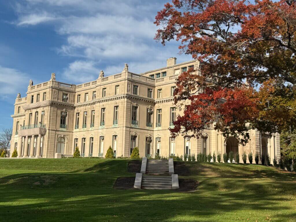 Majestic beige mansion with intricate details stands under a blue sky. In the foreground, a large tree displays vibrant red and orange autumn leaves.