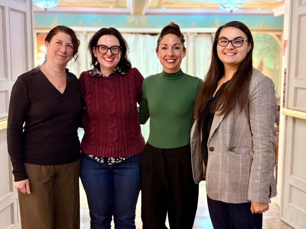 Four women stand together, smiling warmly. They are in a room with elegant decor, featuring soft colors and warm lighting, exuding a friendly atmosphere.