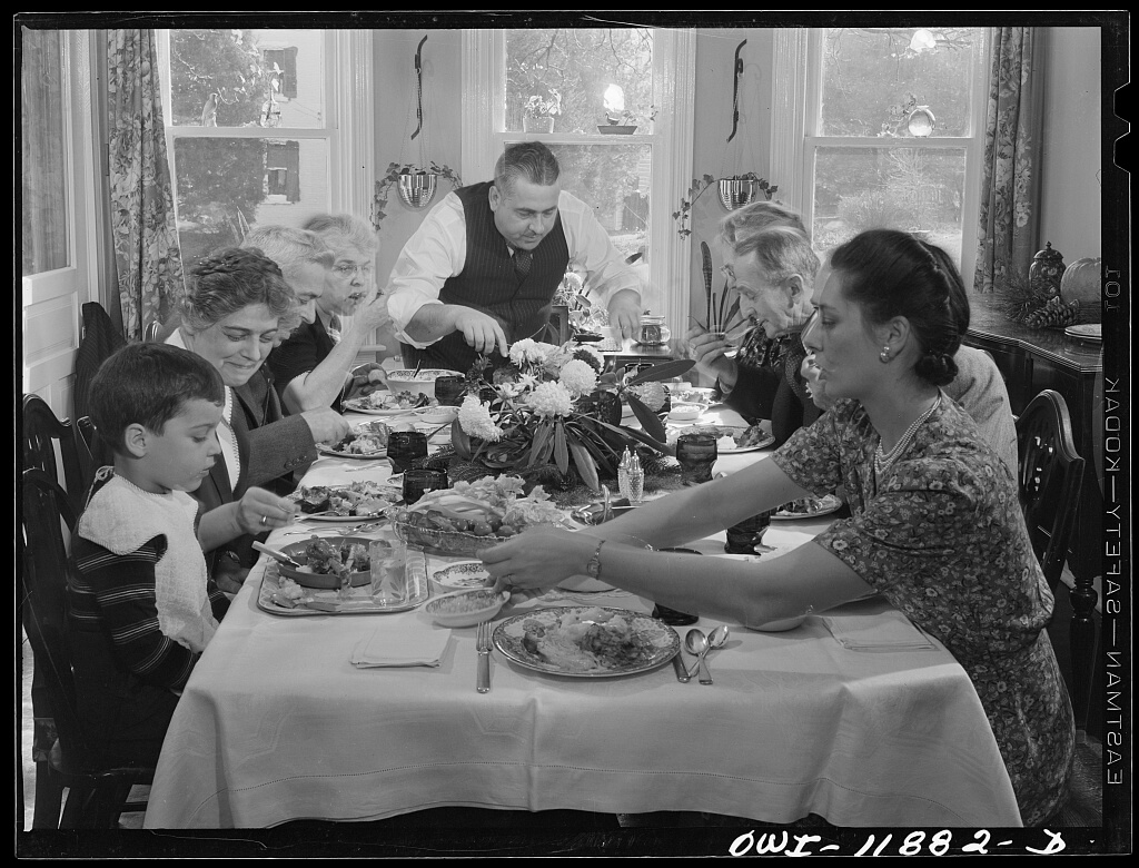 A family gathered around a dining table, enjoying a meal. The table is full of plates with food and a floral centerpiece. The mood is warm and engaged.