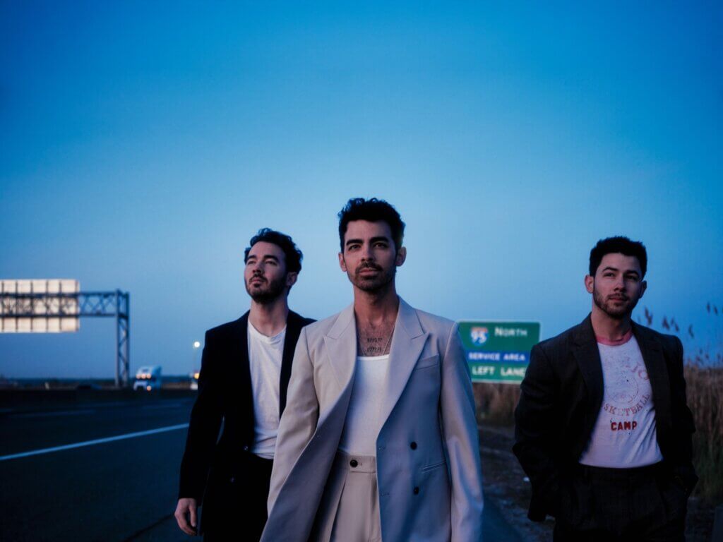Three men walk confidently along a highway at dusk. They wear stylish suits, exuding a cool, determined vibe. A distant road sign and vehicle are visible.