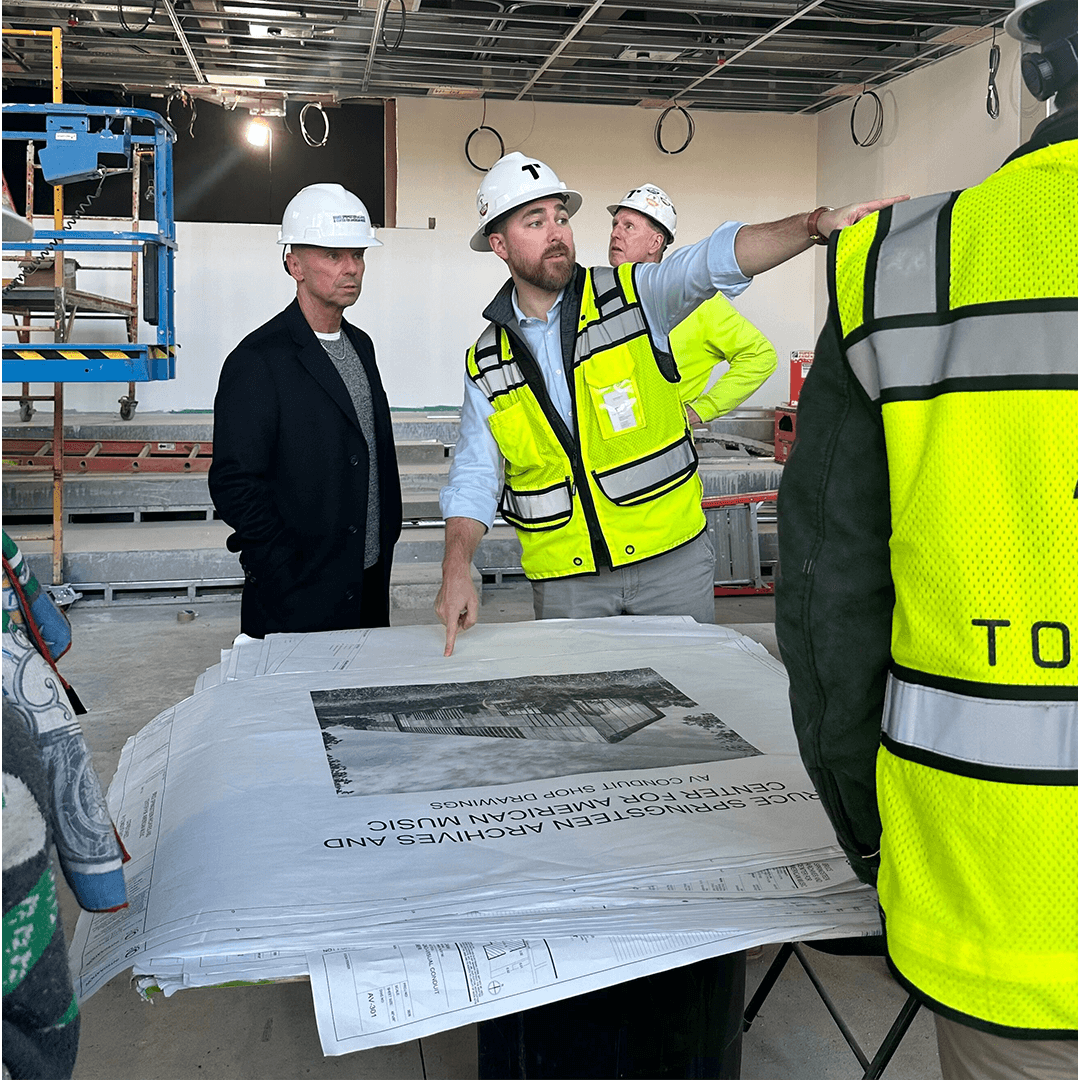 A group of construction workers and a man in a suit discuss architectural blueprints on-site. The man pointing at the plans wears a white hard hat and yellow safety vest, highlighting collaboration and focus in a partially constructed room.