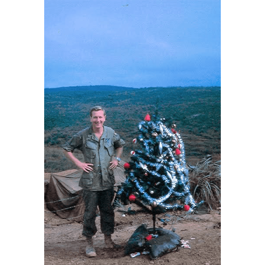 Man in military uniform stands beside a small, decorated Christmas tree outdoors. Hilly landscape in the background under a clear blue sky.