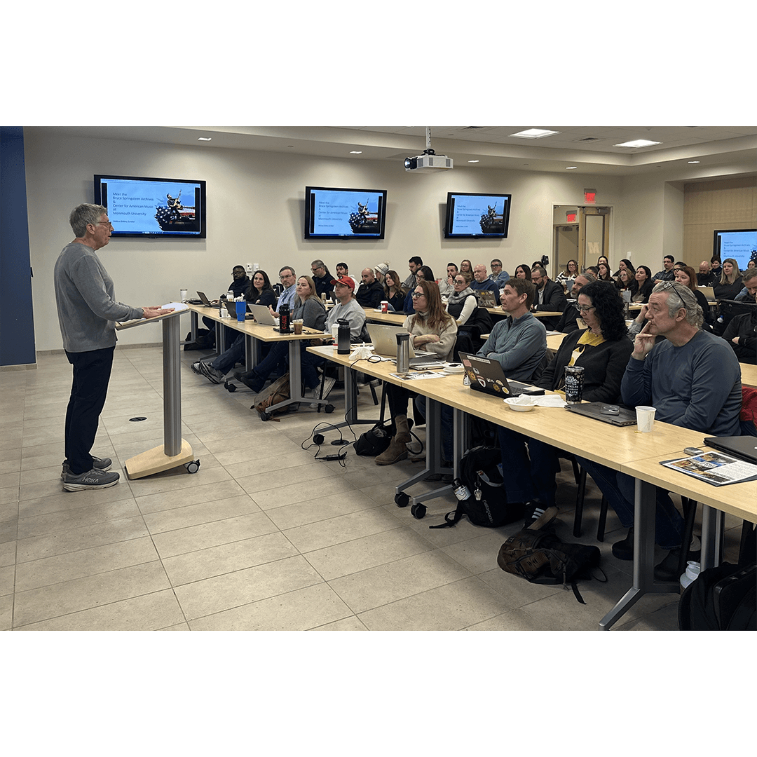 A person is speaking at a podium to a full classroom of attentive adults seated in rows with laptops. The room has multiple wall screens displaying a slide.