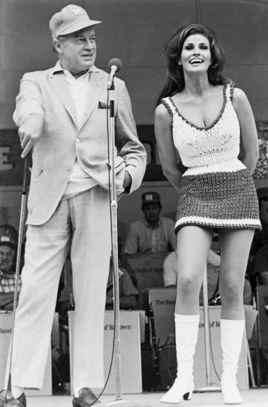 A man in a light suit and hat with a cane stands next to a smiling woman in a crochet mini dress and white boots. They are on stage with a band behind them.