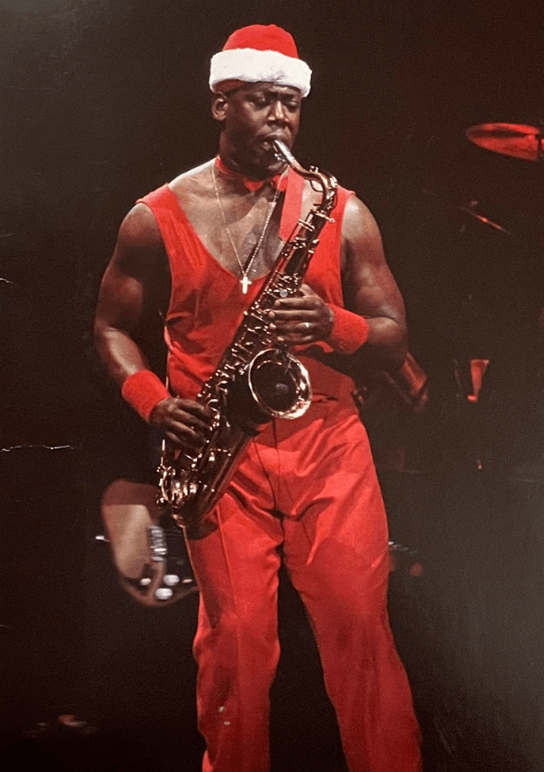 A man in a Santa hat and red outfit plays a saxophone on stage, conveying a festive and energetic mood. The spotlight highlights his concentration.