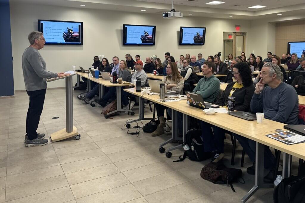 A person is speaking at a podium to a full classroom of attentive adults seated in rows with laptops. The room has multiple wall screens displaying a slide.