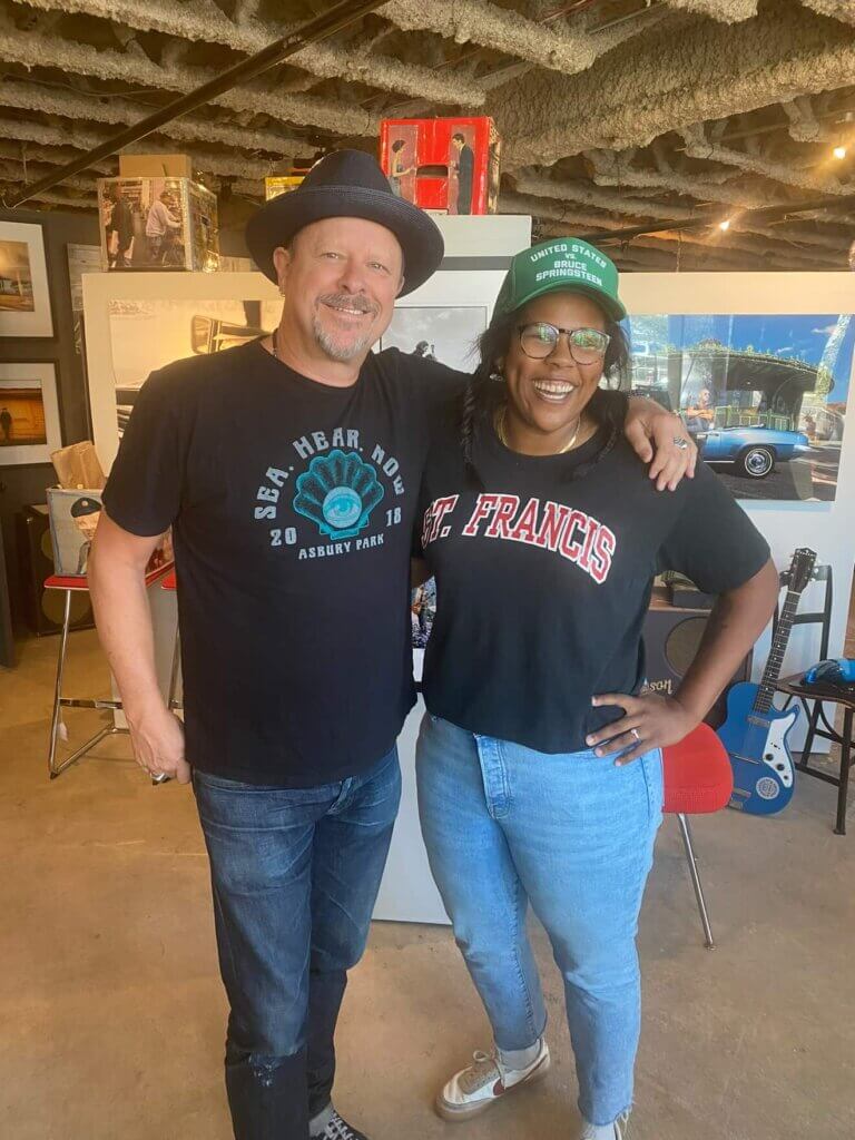 Two people smiling and standing together in a room with guitars and art. One wears a black hat and tee, the other a green cap, black shirt, and jeans.