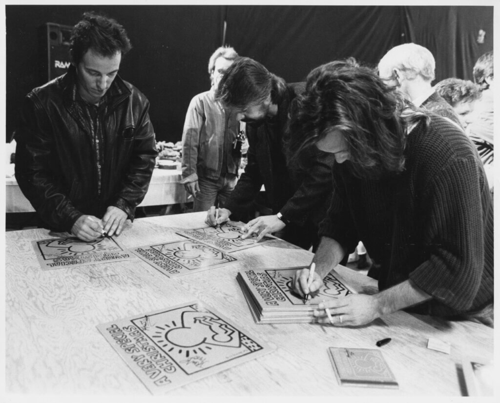 People signing posters on a table covered with Keith Haring designs. The mood is focused, with individuals deeply engaged in their task. Black and white photo.