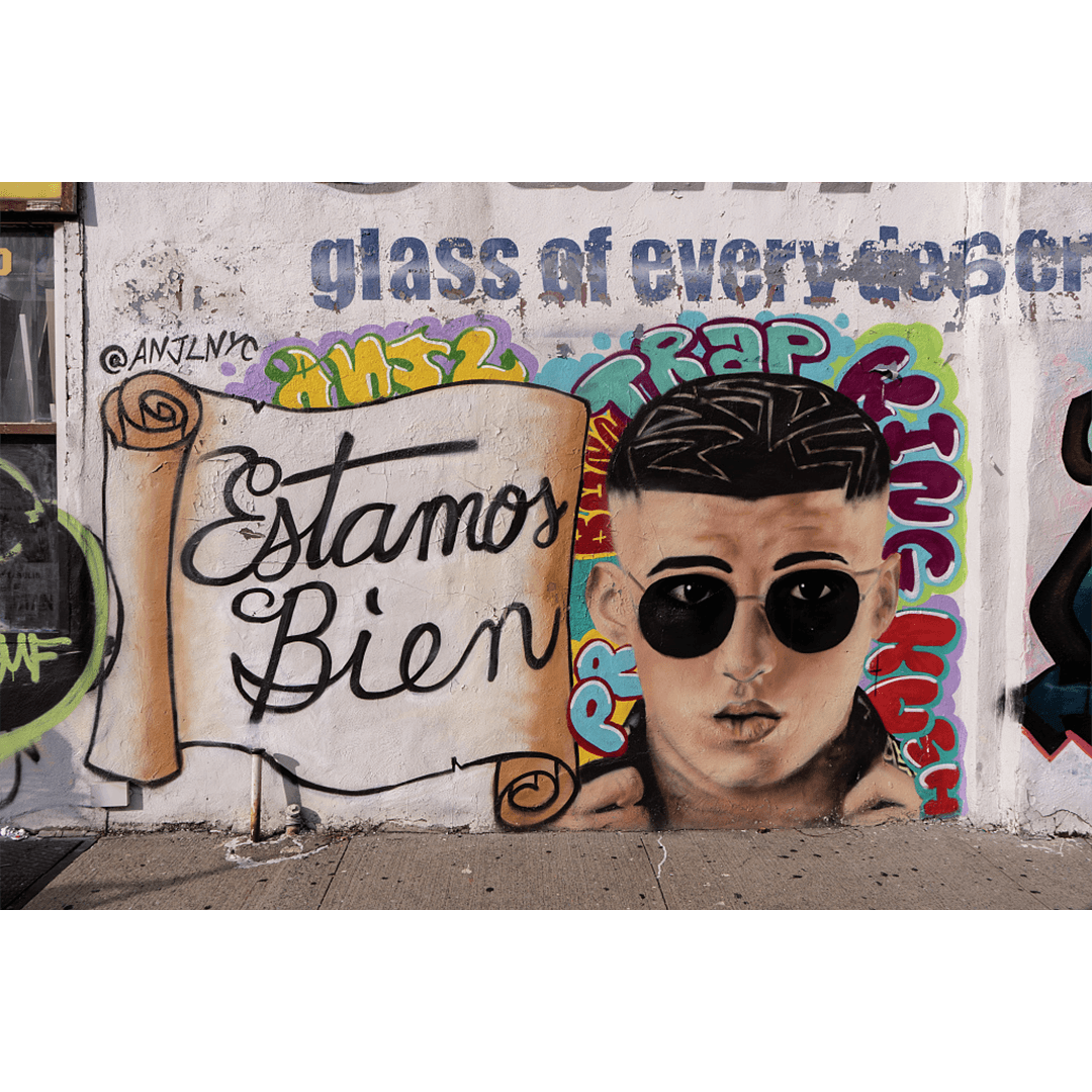 Graffiti mural featuring a scroll with "Estamos Bien" and a stylized portrait of a man in sunglasses. Bright colors and urban street art vibe.