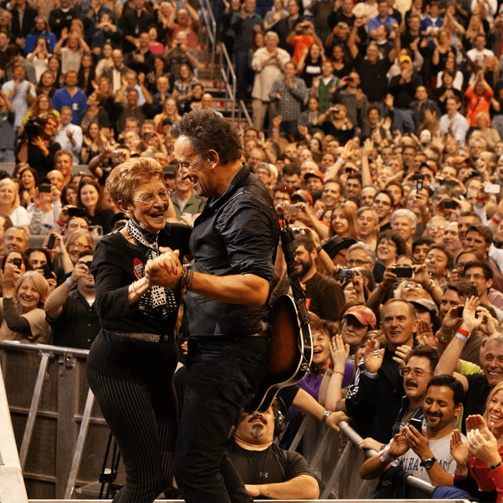 Musician dances joyfully on stage with an elderly woman in front of a cheering concert crowd. The atmosphere is lively and celebratory.