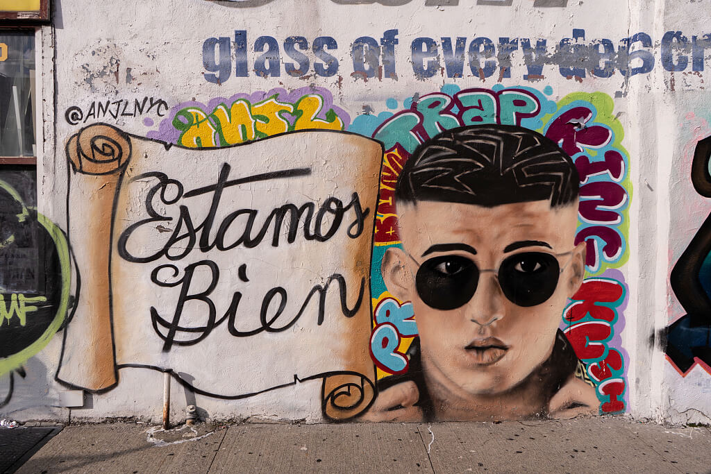 Graffiti mural featuring a scroll with "Estamos Bien" and a stylized portrait of a man in sunglasses. Bright colors and urban street art vibe.