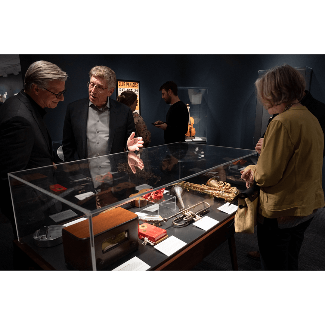 A group of people gathers around a museum display case containing musical instruments and artifacts. The room has a dark, intimate gallery ambiance.