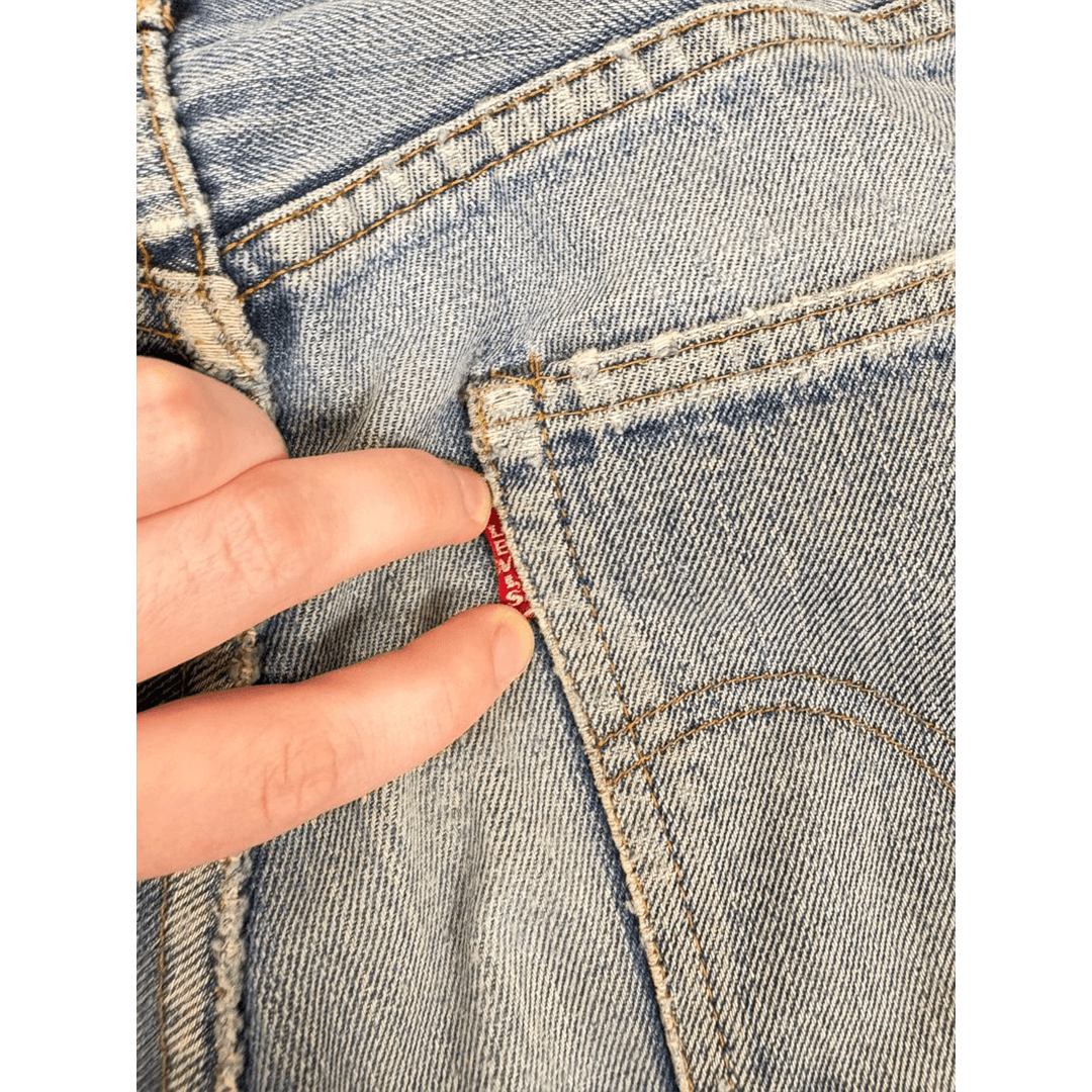 Close-up of a hand holding the small red label of a back pocket on light blue denim jeans. The stitching and fabric texture are clearly visible.