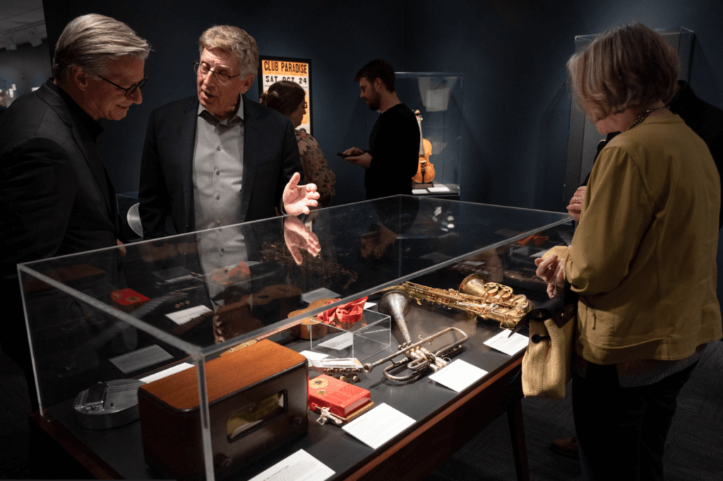 A group of people gathers around a museum display case containing musical instruments and artifacts. The room has a dark, intimate gallery ambiance.