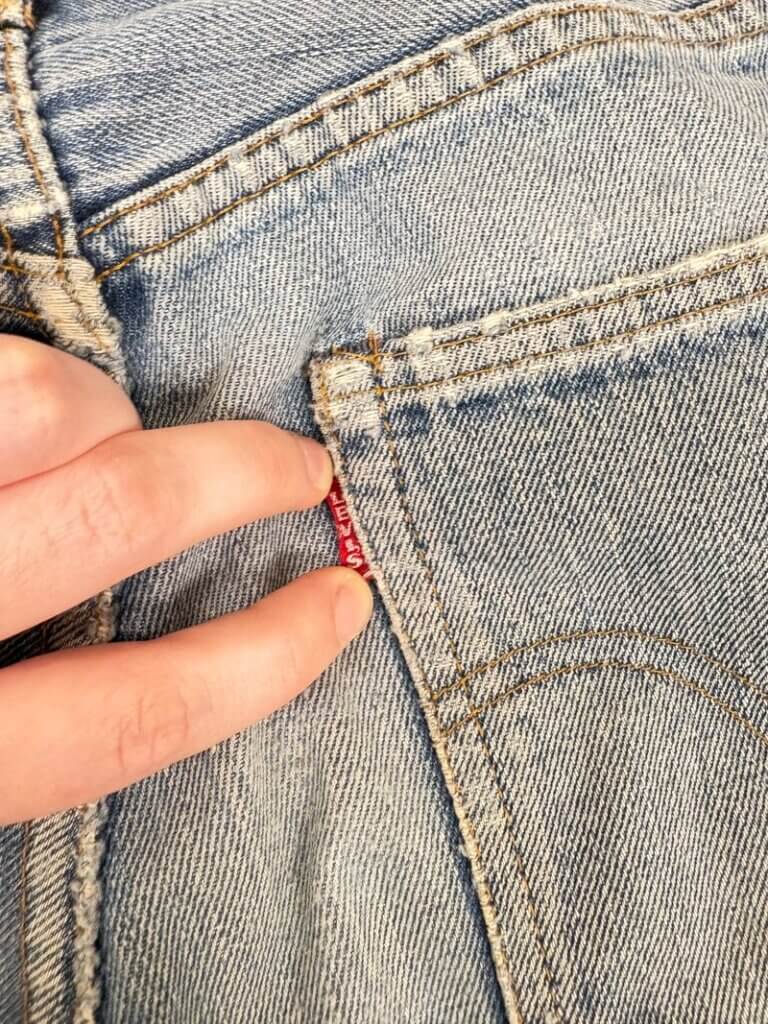 Close-up of a hand holding the small red label of a back pocket on light blue denim jeans. The stitching and fabric texture are clearly visible.