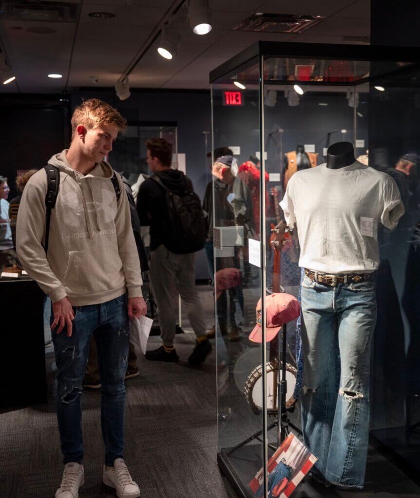 A young man in a white hoodie and jeans observes a museum display case containing a white t-shirt, jeans, a red cap, and a banjo. Other visitors are seen in the background.