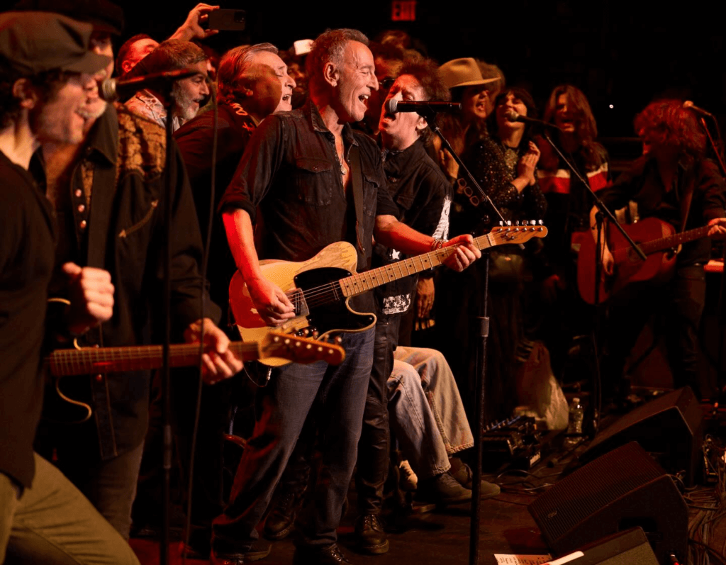 A joyful group of musicians on stage, led by a guitarist in a black shirt, passionately performing. The scene is lively and filled with energy and camaraderie.