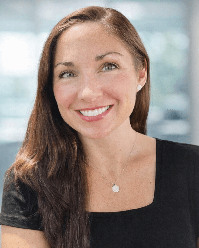 Smiling woman with long brown hair, wearing a black top and a silver necklace, stands in a softly blurred bright setting, conveying warmth and friendliness.