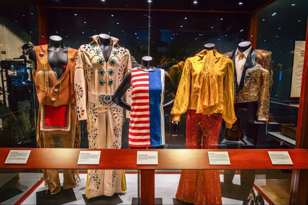 A display of five colorful outfits on mannequins in a museum exhibit, including a white jumpsuit with jewels, a red and blue dress, and a glittering jacket.