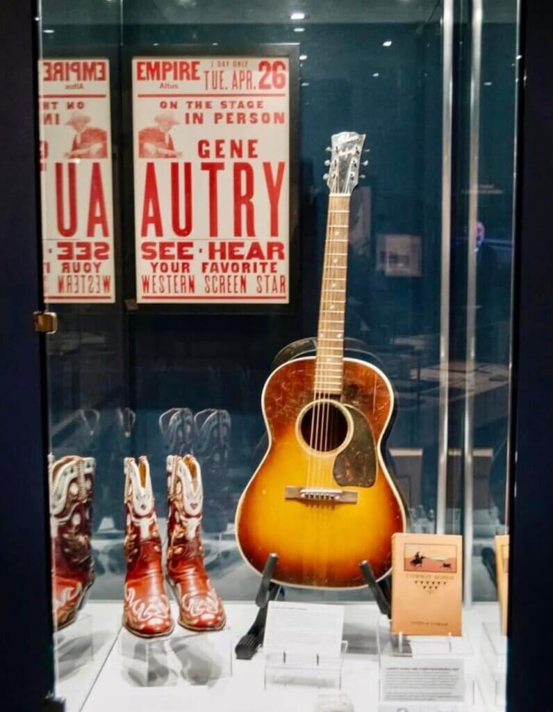 A display case holds a vintage guitar, red cowboy boots, and a concert poster. The items evoke a sense of nostalgia and tribute to classic Western music.