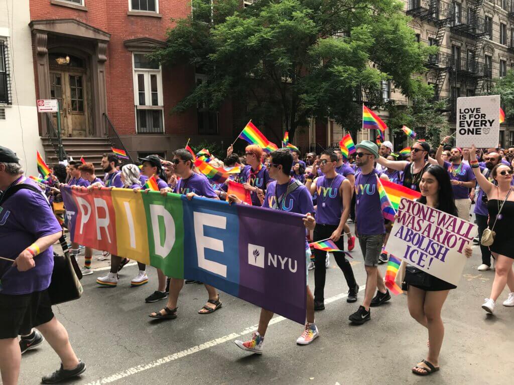 A vibrant Pride parade features individuals in purple shirts holding rainbow flags and a large "PRIDE" banner. Signs advocate for equality and love.