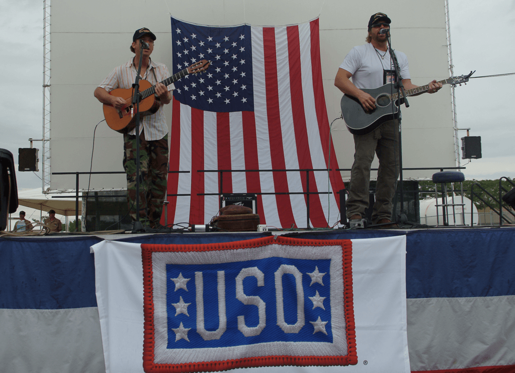Two musicians perform on stage in front of a large American flag, playing guitars. A USO banner hangs below, evoking a patriotic, supportive atmosphere.