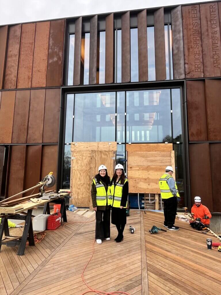 Two people in hard hats and reflective vests stand smiling on a wooden deck in front of an under-construction building. Workers and tools are present.