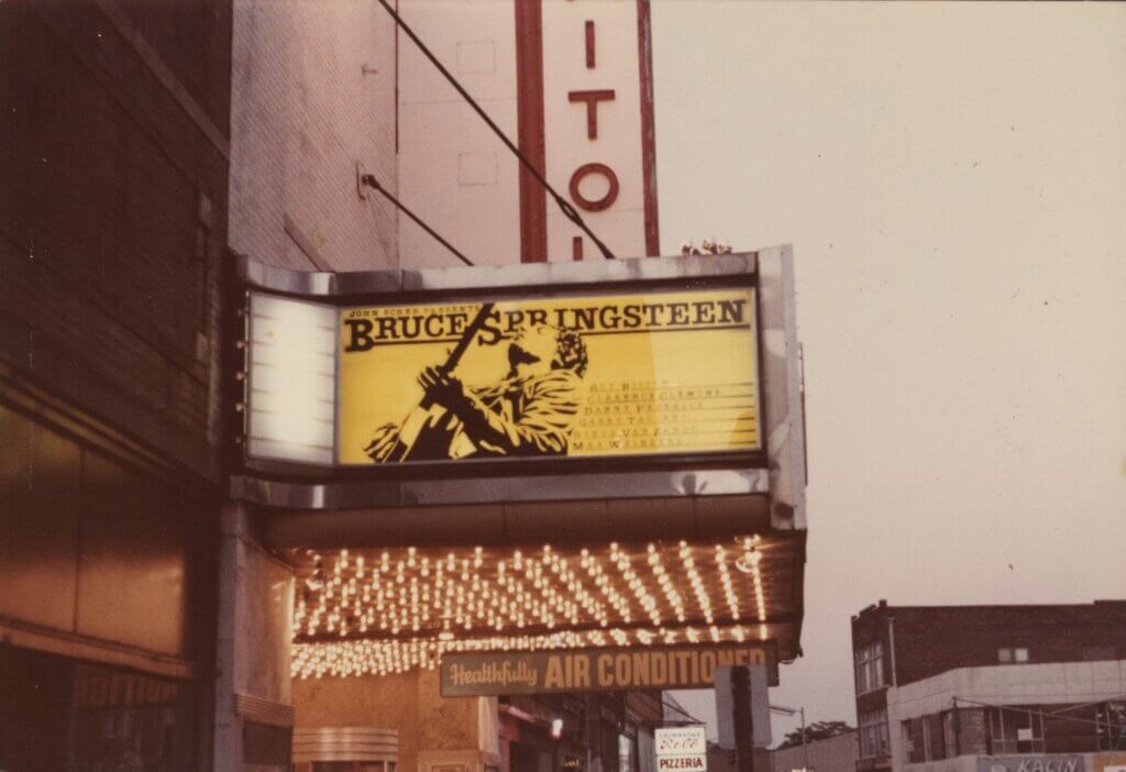 A theater marquee displays "Bruce Springsteen" in bold letters with a musician's silhouette on a guitar against a yellow background, under glowing lights.