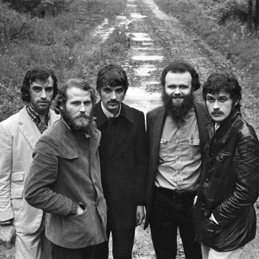 Five men stand closely on a rural dirt road, dressed in vintage 1960s attire. The mood is relaxed and introspective, with trees in the background.