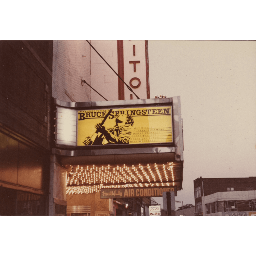 A theater marquee displays "Bruce Springsteen" in bold letters with a musician's silhouette on a guitar against a yellow background, under glowing lights.