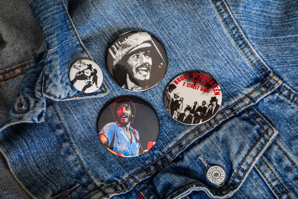 A denim jacket adorned with four buttons. The buttons feature a musician in various expressive poses and a band name, conveying a vintage rock theme.