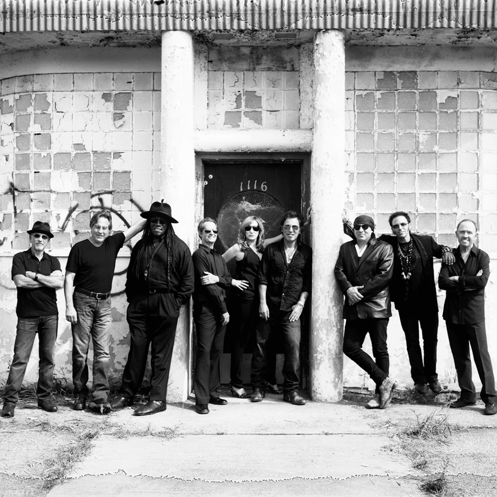 Eight people stand confidently in black clothing against a worn, urban building with peeling paint and graffiti, exuding a cool, relaxed vibe.
