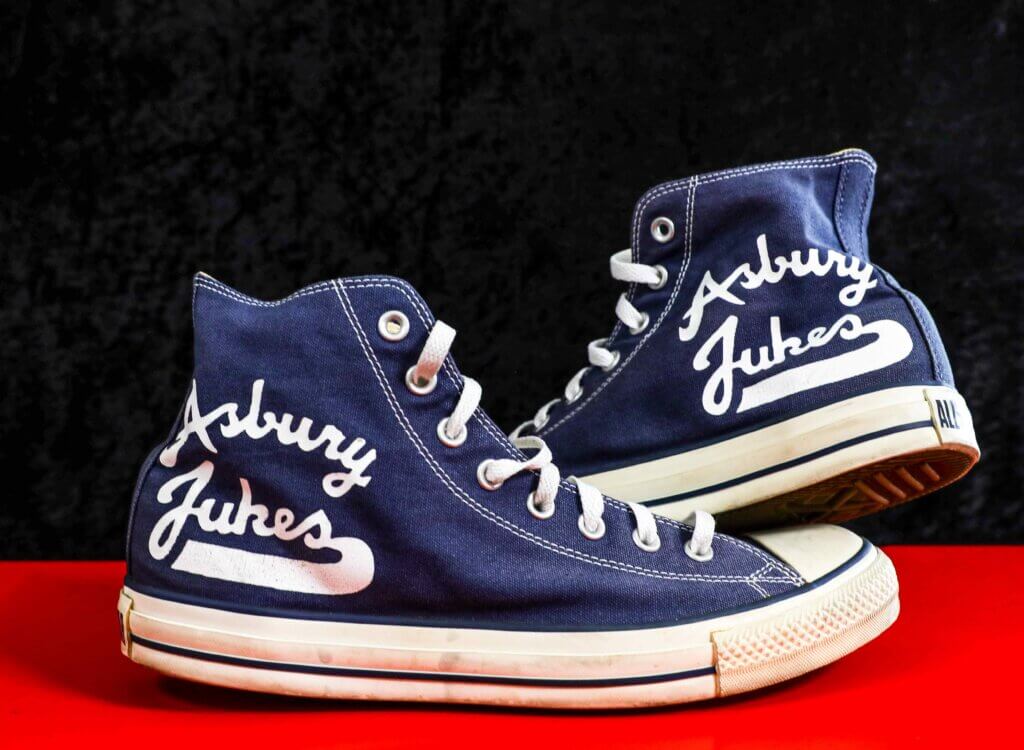 Blue high-top sneakers with "Asbury Jukes" text in white on sides, displayed against a black and red background, conveying a casual, retro vibe.
