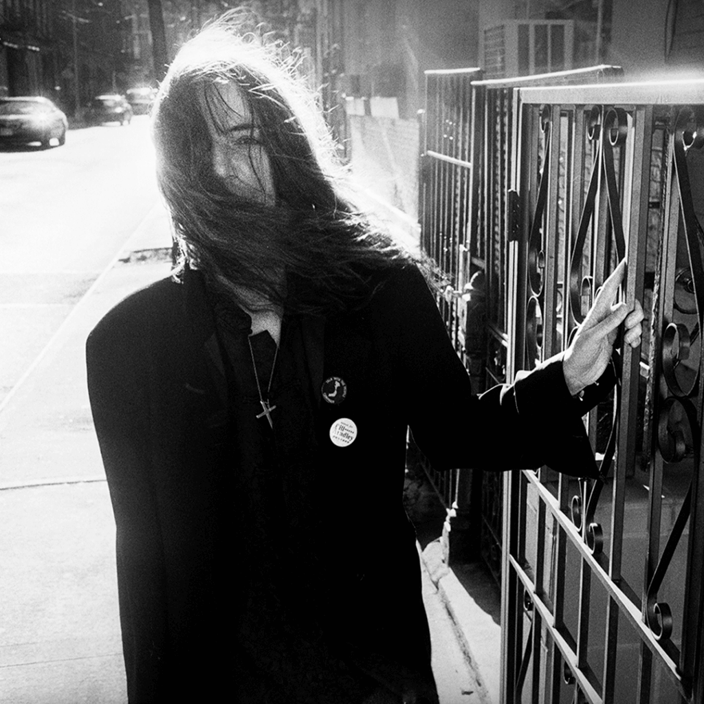 Black and white image of a person on a sunlit street, long hair blown across their face. They wear a coat with badges, creating a mysterious tone.