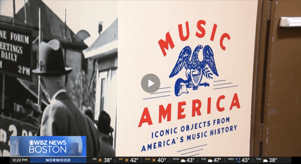 "A poster in a museum setting featuring an eagle with a guitar labeled 'Music America: Iconic Objects from America's Music History.' To the left, a vintage black-and-white photo shows a man in a hat looking at a building. WBZ News Boston ticker at the bottom shows weather and time."