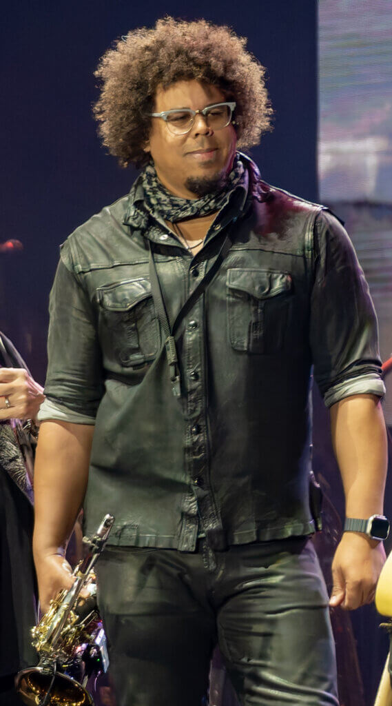 A musician stands confidently on stage, holding a saxophone. He wears a black leather shirt and glasses, with a curly hairstyle and a relaxed expression.