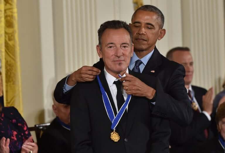 A man places a blue and gold medal around another man's neck in a formal indoor setting, with people seated and applauding in the background.