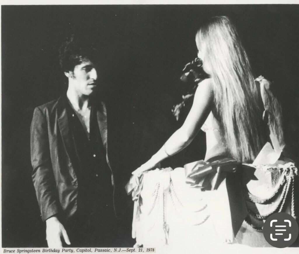 A man in a dark suit gazes at a woman with long hair, seated on a stage decorated with fabric and beads, creating a nostalgic and contemplative mood.